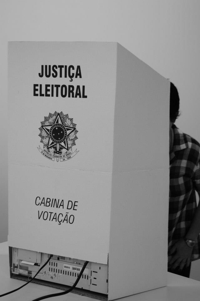 Fotografia em preto e branco de uma cabine de votação da Justiça Eleitoral, com uma pessoa parcialmente visível atrás dela.