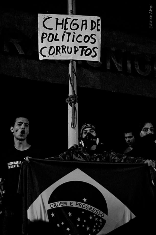 Fotografia em preto e branco de um protesto à noite: um homem de máscara segura a bandeira do Brasil sob um cartaz escrito Chega de políticos corruptos.