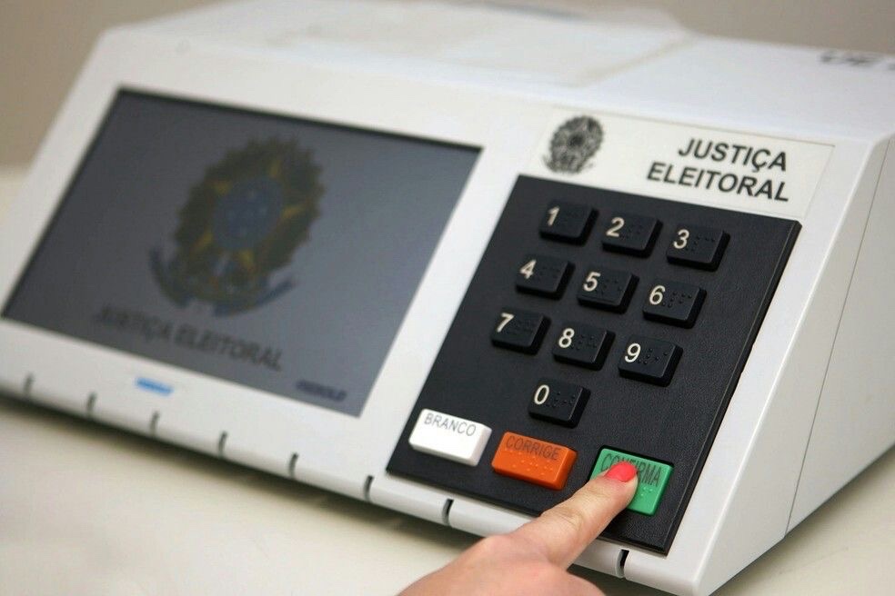 Urna eletrônica da Justiça Eleitoral, com a tela apagada e o dedo de uma pessoa sobre a tecla verde Confirma.